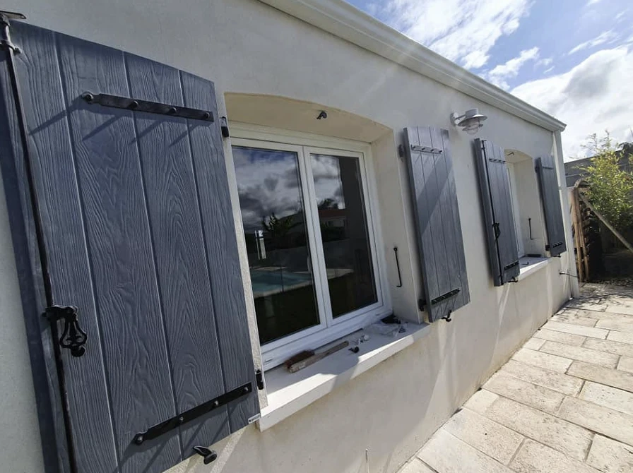 Volets battants veinés gris à pentures et espagnolettes noires, ouverts sur une façade blanche, au soleil
