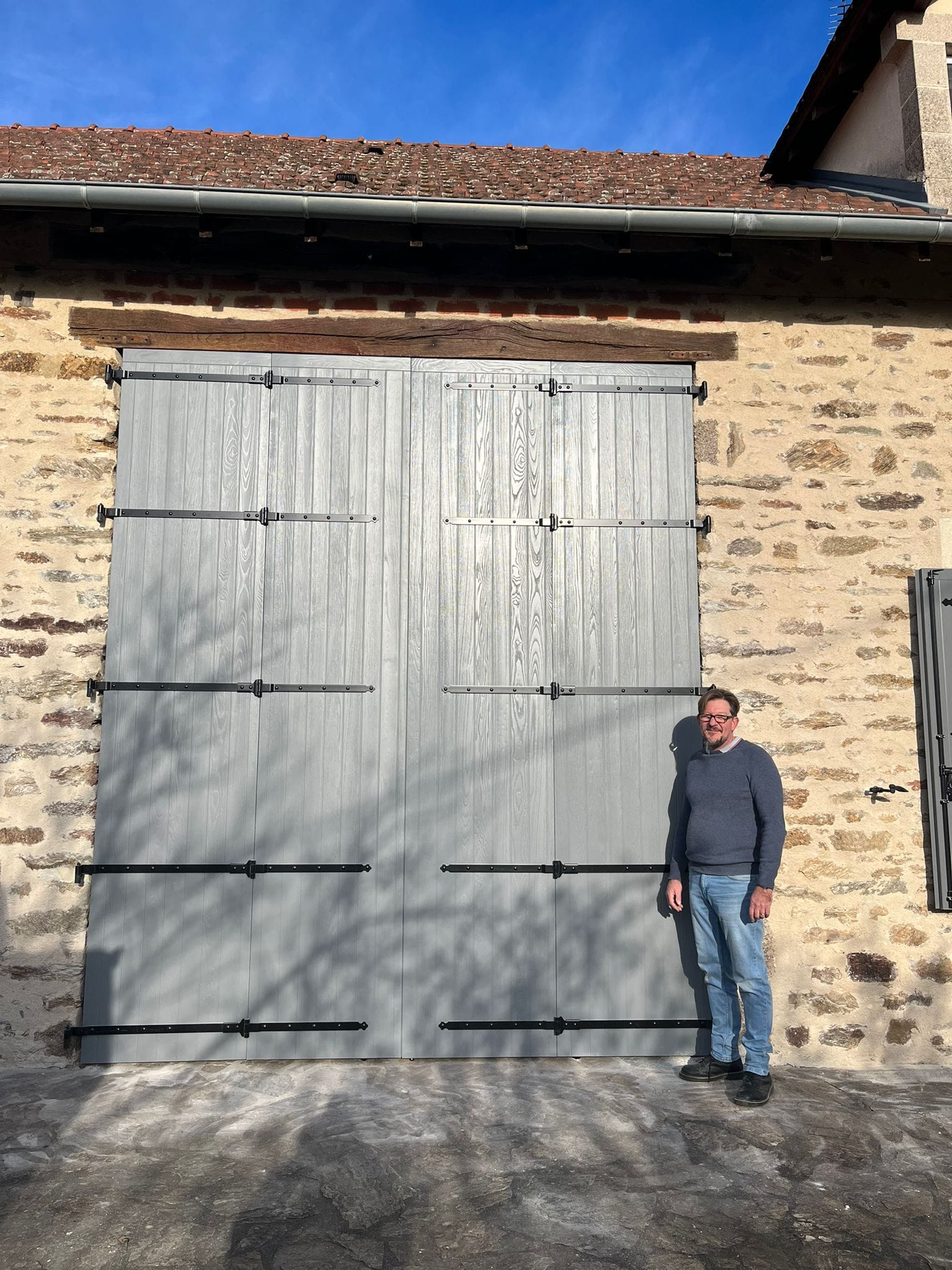 Grands volets battants composites veinés gris à pentures noires sur un bâtiment en pierre, un homme debout à côté
